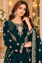 A woman modeling a dark teal green traditional embroidered dress with ornate gold and silver mirror work on the neckline and sleeves, paired with a matching embroidered dupatta, set against a softly blurred indoor background with warm lights and floral arrangements.